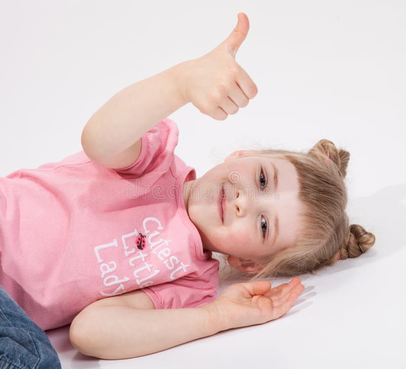 Petite fille de sourire se trouvant sur le plancher et montrant le pouce images libres de droits Petite fille de sourire se trouvant sur le plancher et montrant le pouce images libres de droits