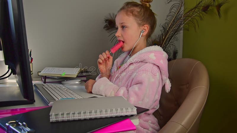 Petite fille en robe de chambre fait les devoirs à l'ordinateur et mange de la crème glacée banque de vidéos