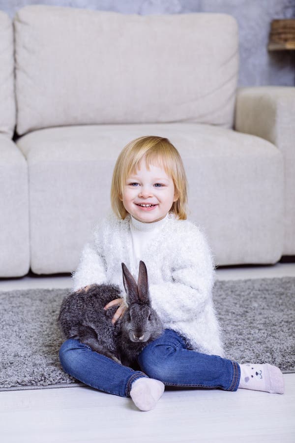 Petite fille mignonne avec le lapin gris de Pâques, lapin Bébé blond souriant dans le chandail blanc et des blues-jean se reposan photographie stock Petite fille mignonne avec le lapin gris de Pâques, lapin Bébé blond souriant dans le chandail blanc et des blues-jean se reposan photographie stock