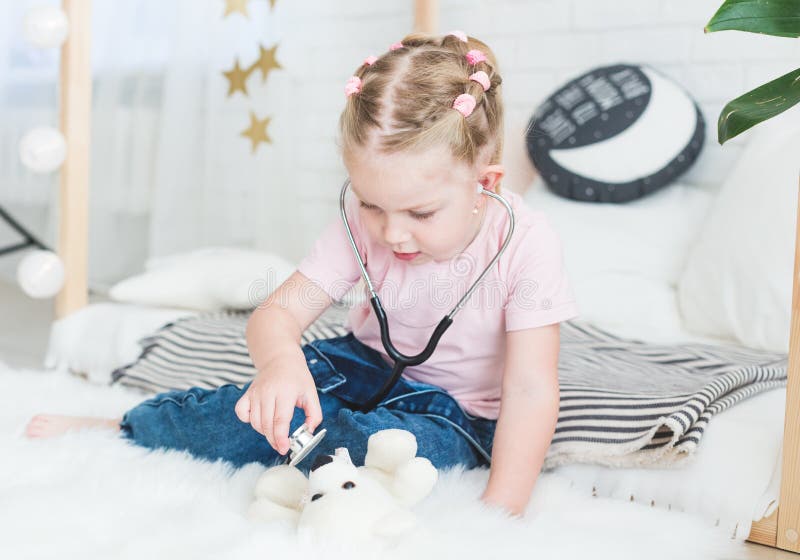 Petite fille mignonne s'asseyant sur le lit et jouant le docteur avec le stéthoscope et le Teddy Bear photo libre de droits Petite fille mignonne s'asseyant sur le lit et jouant le docteur avec le stéthoscope et le Teddy Bear photo libre de droits