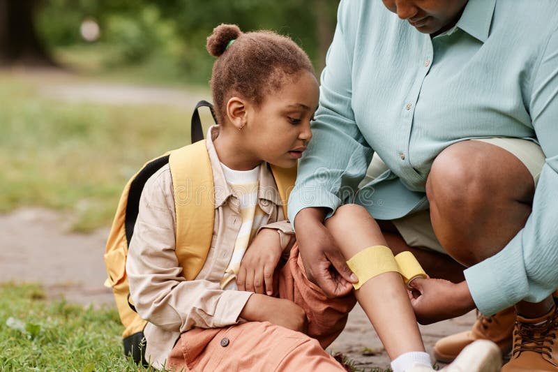 Petite fille noire blessée randonnée avec mère mettre un bandage sur le genou à l'extérieur photo stock