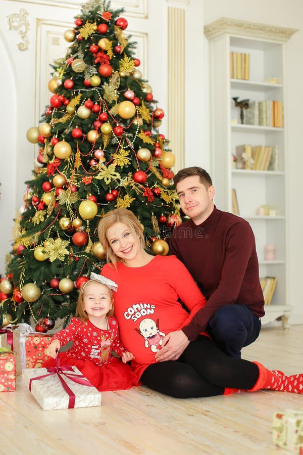 Petite fille portant la robe rouge se reposant avec le père et la mère enceinte près de l'arbre de Noël et gardant des cadeaux images stock Petite fille portant la robe rouge se reposant avec le père et la mère enceinte près de l'arbre de Noël et gardant des cadeaux images stock