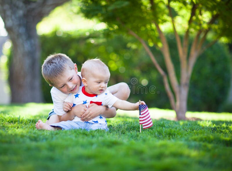 Petits frères mignons s'asseyant sur une pelouse verte tenant le drapeau américain photos stock Petits frères mignons s'asseyant sur une pelouse verte tenant le drapeau américain photos stock