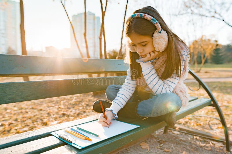Peu beau dessin d'artiste avec les crayons colorés, fille s'asseyant sur un banc en parc ensoleillé d'automne, heure d'or image libre de droits Peu beau dessin d'artiste avec les crayons colorés, fille s'asseyant sur un banc en parc ensoleillé d'automne, heure d'or image libre de droits