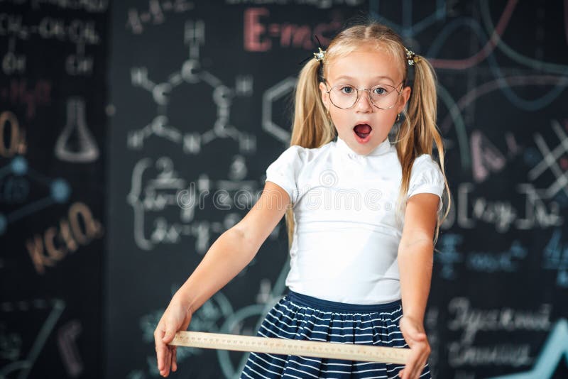 Peu fille blonde avec un visage étonné avec une règle dans sa fille d'élève de mains avec de grandes règles contre le tableau ave images libres de droits Peu fille blonde avec un visage étonné avec une règle dans sa fille d'élève de mains avec de grandes règles contre le tableau ave images libres de droits