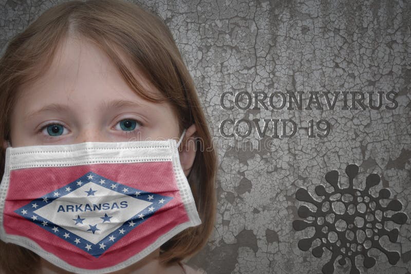 Little girl in medical mask with arkansas state flag stands near the old vintage wall with text coronavirus, covid, and virus picture. Stop virus. Little girl in medical mask with arkansas state flag stands near the old vintage wall with text coronavirus, covid, and virus picture. Stop virus