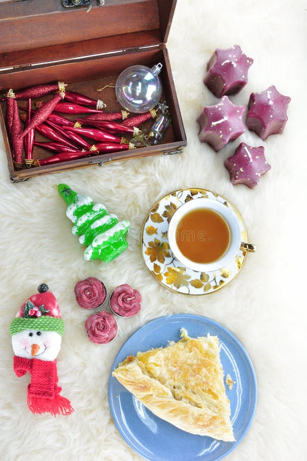 Piece of greek feta cheese pie, a tea cup with matching saucer, a wooden box with Christmas ornaments, tree shaped,rose shape and star shaped candles and a snowman face ornament on sheepskin carpet. Piece of greek feta cheese pie, a tea cup with matching saucer, a wooden box with Christmas ornaments, tree shaped,rose shape and star shaped candles and a snowman face ornament on sheepskin carpet