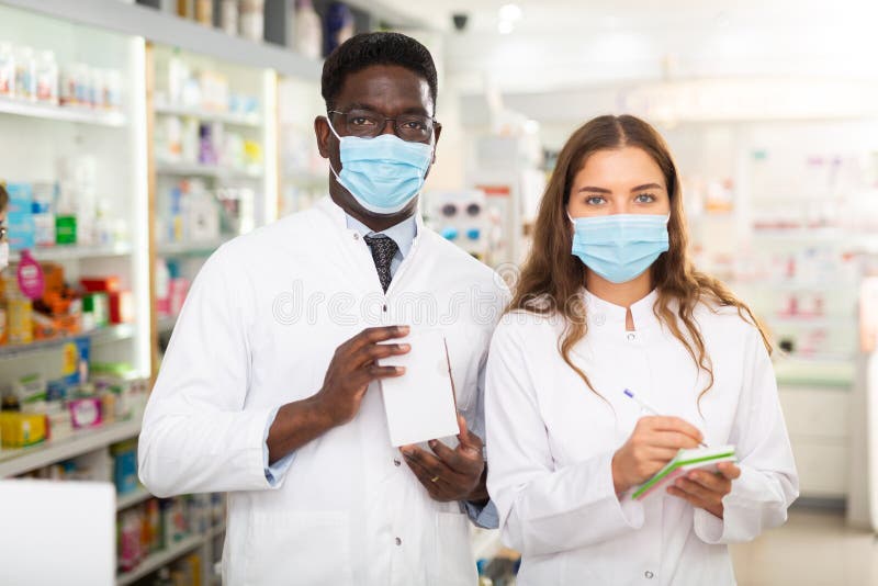 Pharmacists in face masks and white gowns standing in salesroom of drugstore. Pharmacists in face masks and white gowns standing in salesroom of drugstore