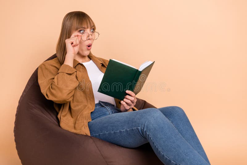 Photo of astonished shocked young woman hold glasses book sit bean bag isolated on pastel beige color background stock image