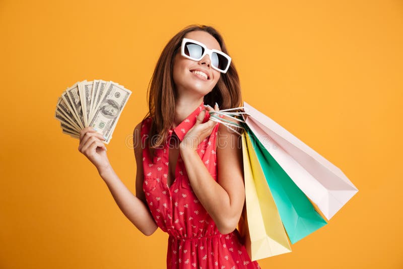 Photo of cheerful elegant young woman in sunglasses and red dress holding fan of money and colorful shopping bags, looking aside