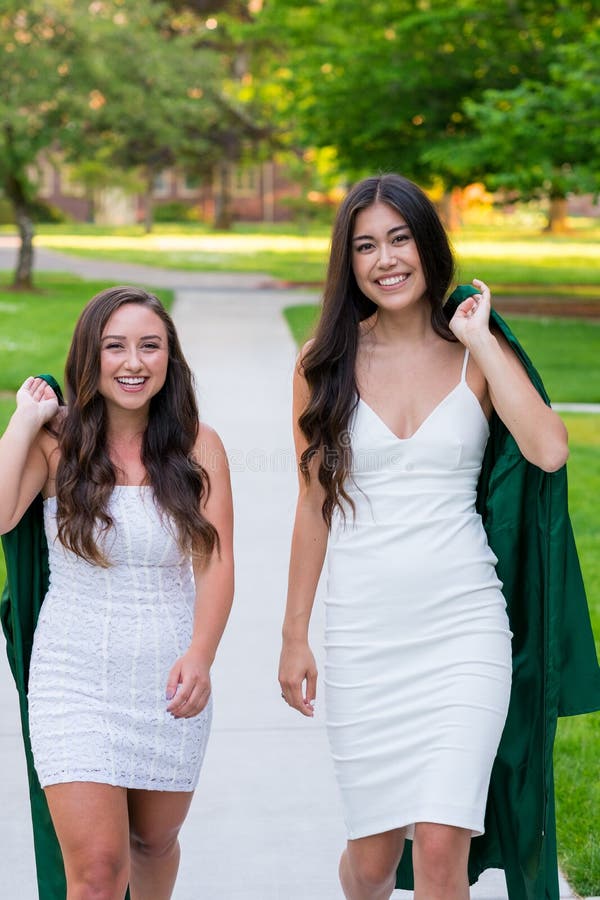 Two college girls and best friends walk on a sidewalk while holding their caps and gowns before graduation on a university campus during the Spring in Oregon. Two college girls and best friends walk on a sidewalk while holding their caps and gowns before graduation on a university campus during the Spring in Oregon.