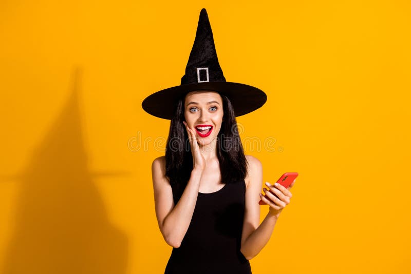 Photo of pretty lovely young magician lady hold telephone hand, cheek open mouth excited find online shop discount wear black wizard headwear dress isolated bright yellow color background. Photo of pretty lovely young magician lady hold telephone hand, cheek open mouth excited find online shop discount wear black wizard headwear dress isolated bright yellow color background