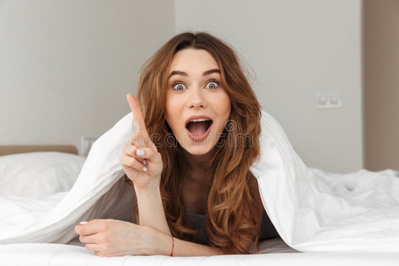 Photo of excited woman 20s lying in bed under white blanket in bedroom and having idea putting index finger up