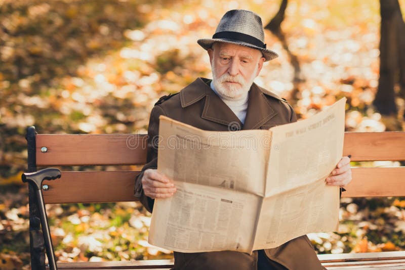 Photo of focused old man grandfather sit bench autumn town park read interesting bought newspaper wear cap headwear