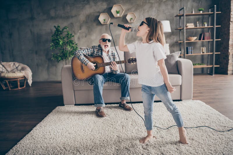 Photo of grandpa sit sofa play guitar little pretty granddaughter hold mic singing song spend time together cool trendy, sun glasses stay home quarantine rejoicing modern living room indoors