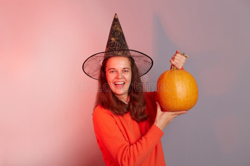 Indoor shot of smiling excited overjoyed young happy woman in black witch hat showing ripe pumpkin to camera, celebrating halloween, posing isolated on colorful neon light background. Indoor shot of smiling excited overjoyed young happy woman in black witch hat showing ripe pumpkin to camera, celebrating halloween, posing isolated on colorful neon light background