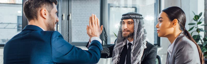 Panoramic shot of professional multicultural business partners giving highfive on meeting with translator in office. Panoramic shot of professional multicultural business partners giving highfive on meeting with translator in office