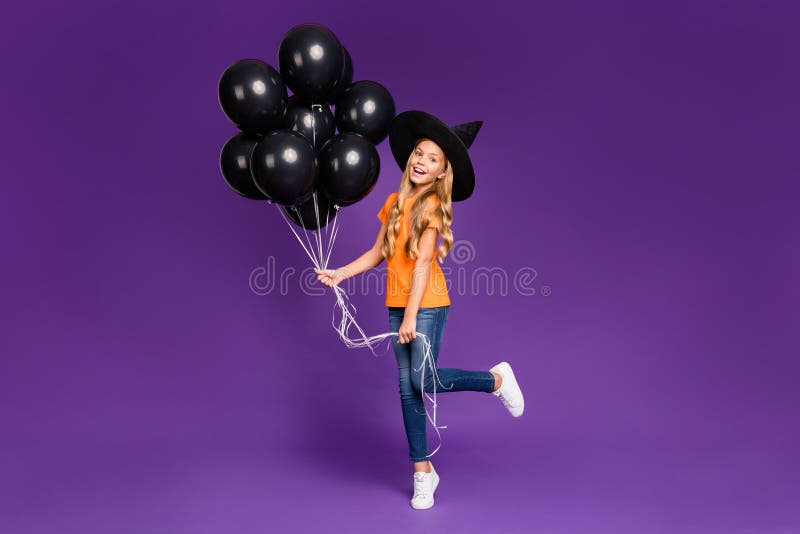 Full size photo of little witch lady halloween party holding many air balloons, excited chilling wear orange t-shirt wizard hat isolated purple color background. Full size photo of little witch lady halloween party holding many air balloons, excited chilling wear orange t-shirt wizard hat isolated purple color background