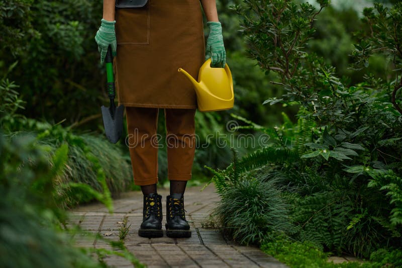 Cropped shot of female gardener dressed in apron holding garden tool in hands wearing rubber gloves view on leg in boots over greenhouse background. Cropped shot of female gardener dressed in apron holding garden tool in hands wearing rubber gloves view on leg in boots over greenhouse background