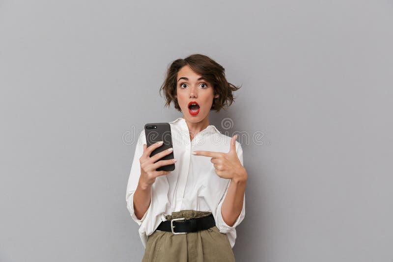 Photo of shocked woman 20s holding and using mobile phone, isolated over gray background