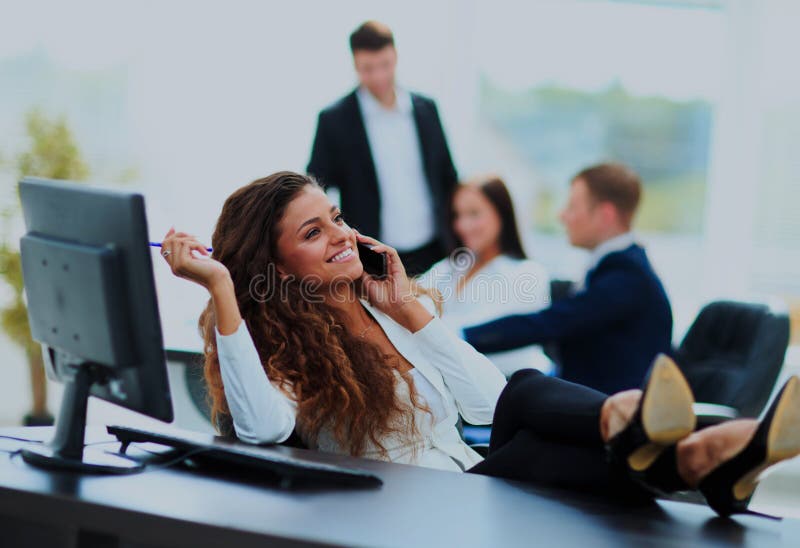 Photo of smiling brunette businesswoman with legs on the desk phoning in office. Photo of smiling brunette businesswoman with legs on the desk phoning in office