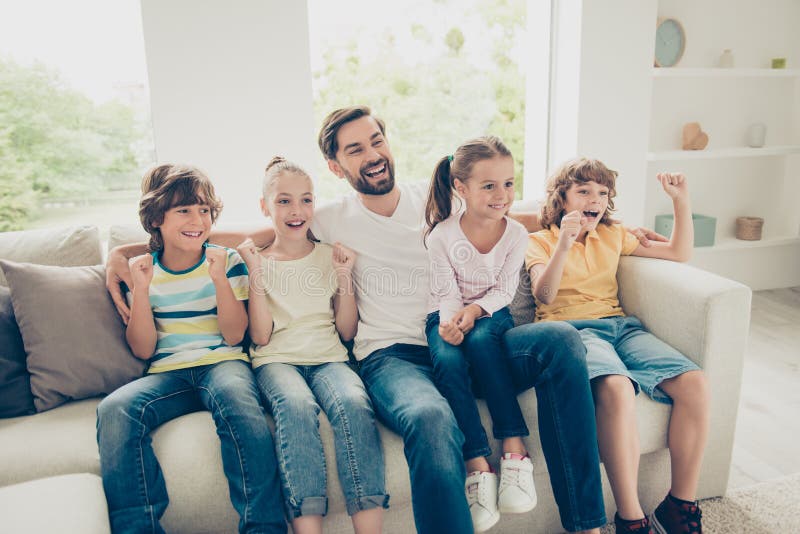 Photo of young father with youngster, youth funky kids big toothy smile, raised hands up sit on comfort, cozy sofa in bright lovely living flat room and look TV