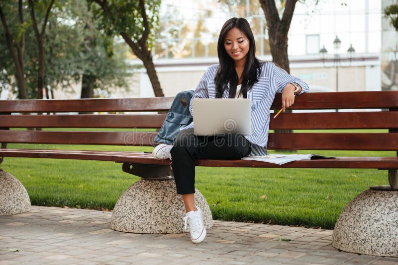 Photo of young happy asian female student relaxing with laptop, sitting on wooden bench in park