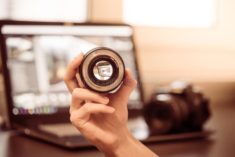 Photographer is holding a photography lens in his hand, camera and laptop in the blurry background