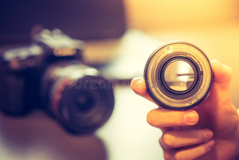 Photographer is holding a photography lens in his hand, camera and laptop in the blurry background