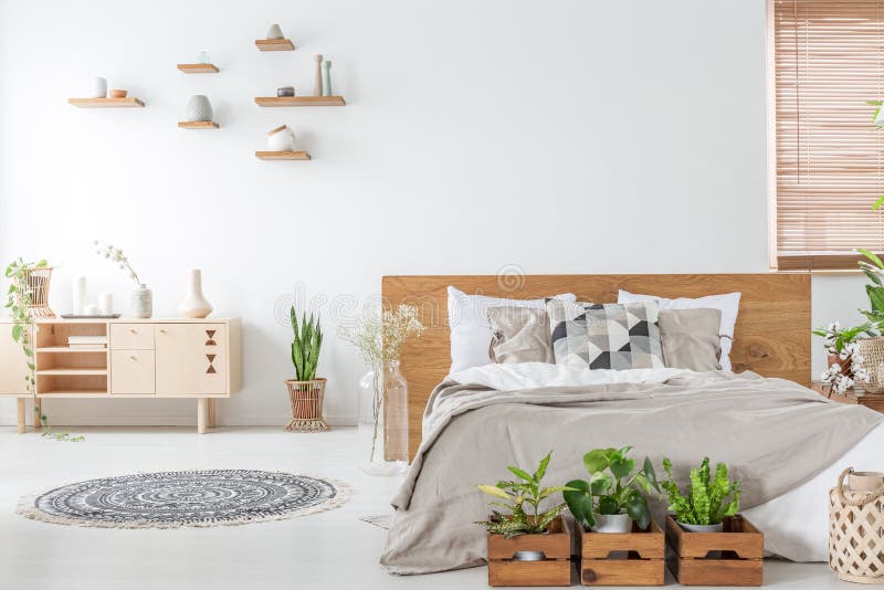 Piante davanti al letto di legno nell'interno bianco della camera da letto con la coperta vicino all'armadietto Foto reale fotografia stock Piante davanti al letto di legno nell'interno bianco della camera da letto con la coperta vicino all'armadietto Foto reale fotografia stock