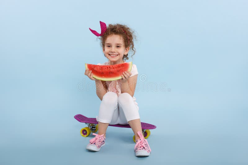 Piccola ragazza dai capelli riccia di felicità con l'anguria in mani, messe su un pattino, isolato su un fondo blu fotografie stock libere da diritti Piccola ragazza dai capelli riccia di felicità con l'anguria in mani, messe su un pattino, isolato su un fondo blu fotografie stock libere da diritti