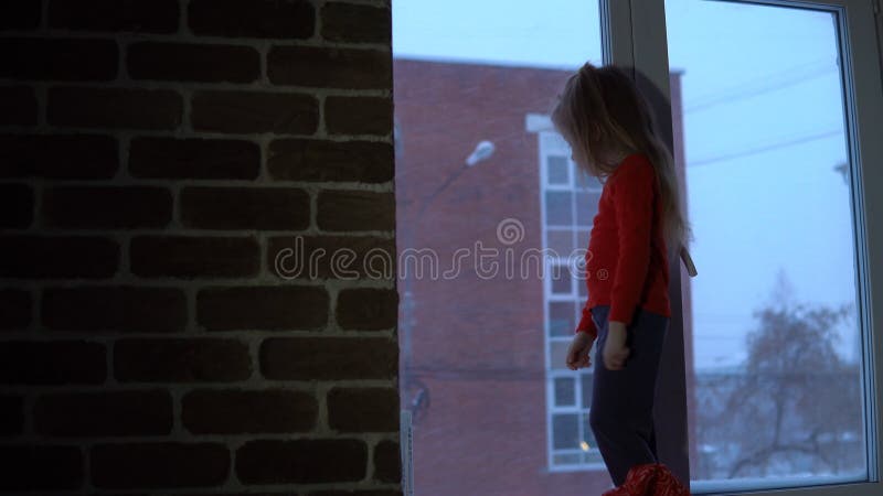 Piccola ragazza sveglia che sta sul davanzale della finestra, guardante fuori su un paesaggio urbano nevoso