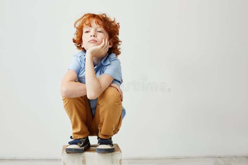 Piccola seduta divertente del bambino della testarossa, testa della tenuta con la mano, guardando da parte con l'espressione vaga fotografia stock libera da diritti
