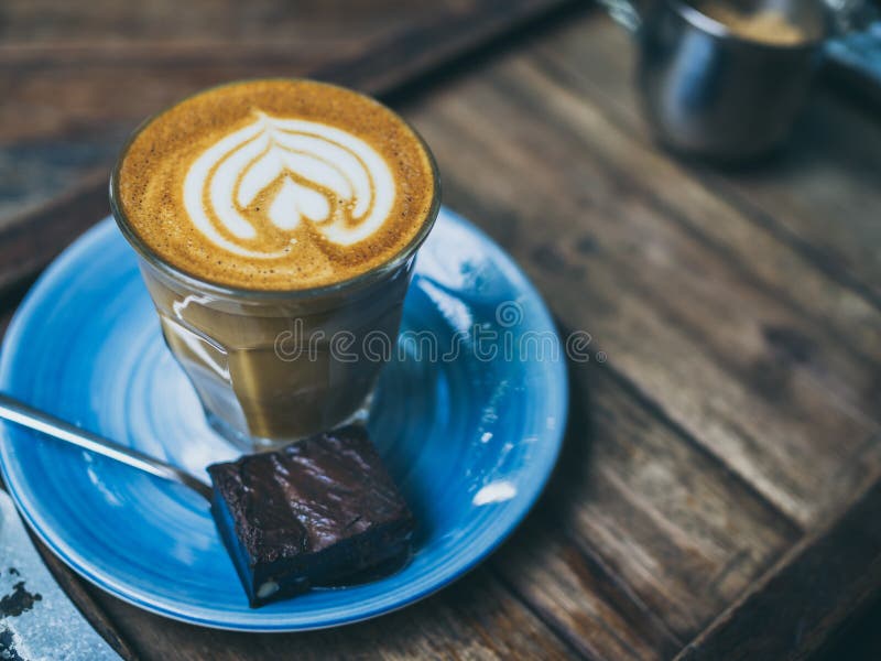 Piccolo Latte coffee topping with flower art from milk in small glass with a piece of homemade brownies cake on blue ceramic plate on wooden tray on wooden table.