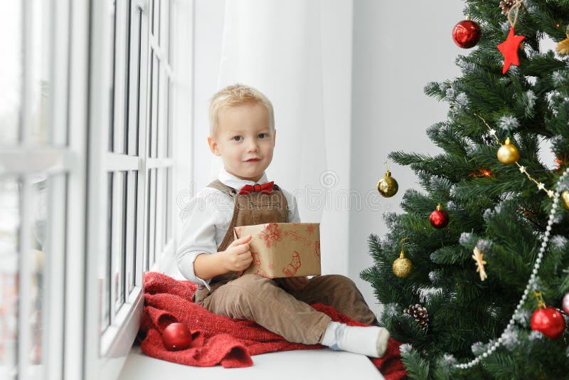 Piccolo neonato con il presente in sue mani che si siedono vicino all'albero di Natale e che esaminano macchina fotografica immagini stock libere da diritti Piccolo neonato con il presente in sue mani che si siedono vicino all'albero di Natale e che esaminano macchina fotografica immagini stock libere da diritti