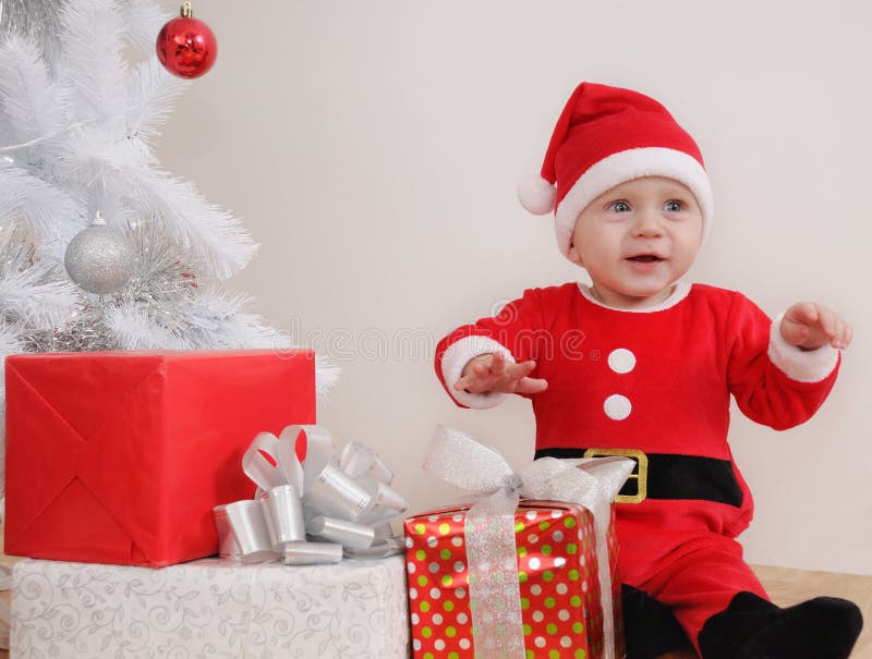 Piccolo neonato felice sveglio nel vestito di Santa con i regali vicino a Christm immagini stock Piccolo neonato felice sveglio nel vestito di Santa con i regali vicino a Christm immagini stock