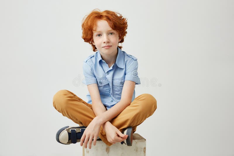 Piccolo ragazzo divertente con capelli arancio e le lentiggini che si siedono sulla scatola di legno in studio, guardando in came immagine stock