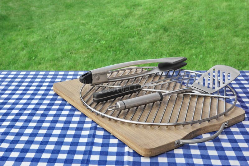 Picnic Table With Checkered Tablecloth, Cutting Board, Grill And Barbecue Tools, Green Lawn In The Background, Summer Outdoor BBQ Party Or Picnic Concept. Picnic Table With Checkered Tablecloth, Cutting Board, Grill And Barbecue Tools, Green Lawn In The Background, Summer Outdoor BBQ Party Or Picnic Concept