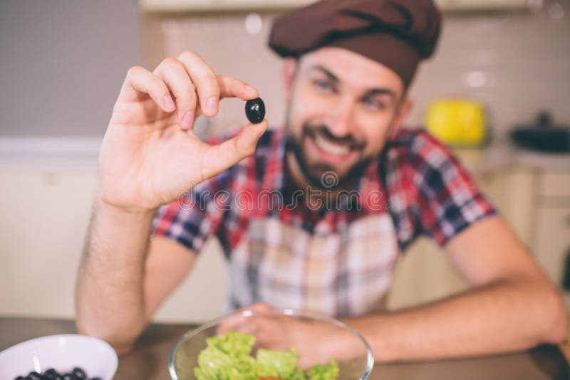 A picture of positive and bearded man leaning on table and hold one piece of black olive. He looks at it. Young man is royalty free stock photo A picture of positive and bearded man leaning on table and hold one piece of black olive. He looks at it. Young man is royalty free stock photo