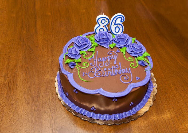 Pictured is a lovely Happy 86th birthday cake with chocolate icing and purple flowers and number candles.  It sits on a brown wooden table top.