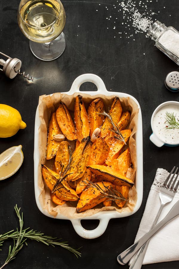 Roasted sweet potatoes in white ceramic dish. Wine, dip, lemon, salt and rosemary around. Black chalkboard as background. Restaurant table scenery from above. Roasted sweet potatoes in white ceramic dish. Wine, dip, lemon, salt and rosemary around. Black chalkboard as background. Restaurant table scenery from above.