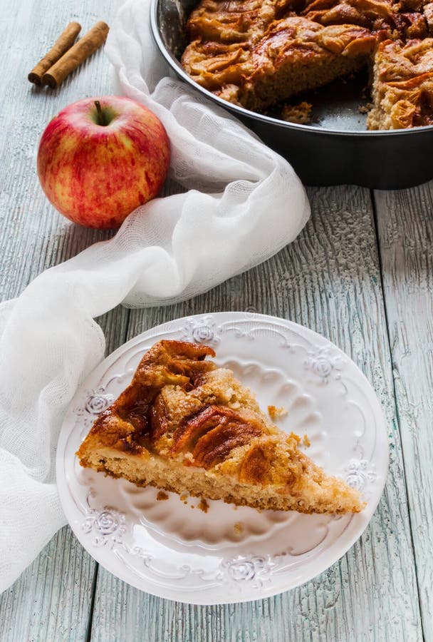 Piece of homemade sweet apple and cinnamon cake on vintage white plate. Pie in baking dish and fresh fruit on wooden background stock photo Piece of homemade sweet apple and cinnamon cake on vintage white plate. Pie in baking dish and fresh fruit on wooden background stock photo