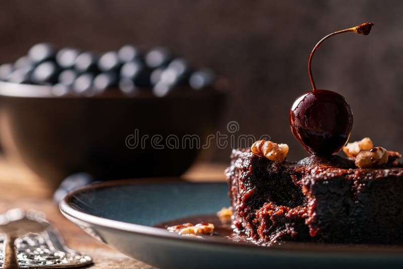 Piece of a homemade vegan chocolate brownie, bitten off and decorated with a cherry. On a plate in front of a bowl of blueberries. Head on view, macro closeup stock photography