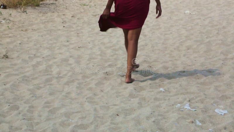 Pieds féminins sur le sable dans une robe rouge en développement. un banque de vidéos