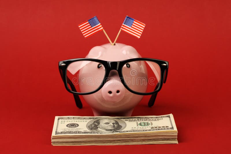 Piggy bank with black spectacle frame of glasses and two small USA flags standing on stack of money american hundred dollar bills