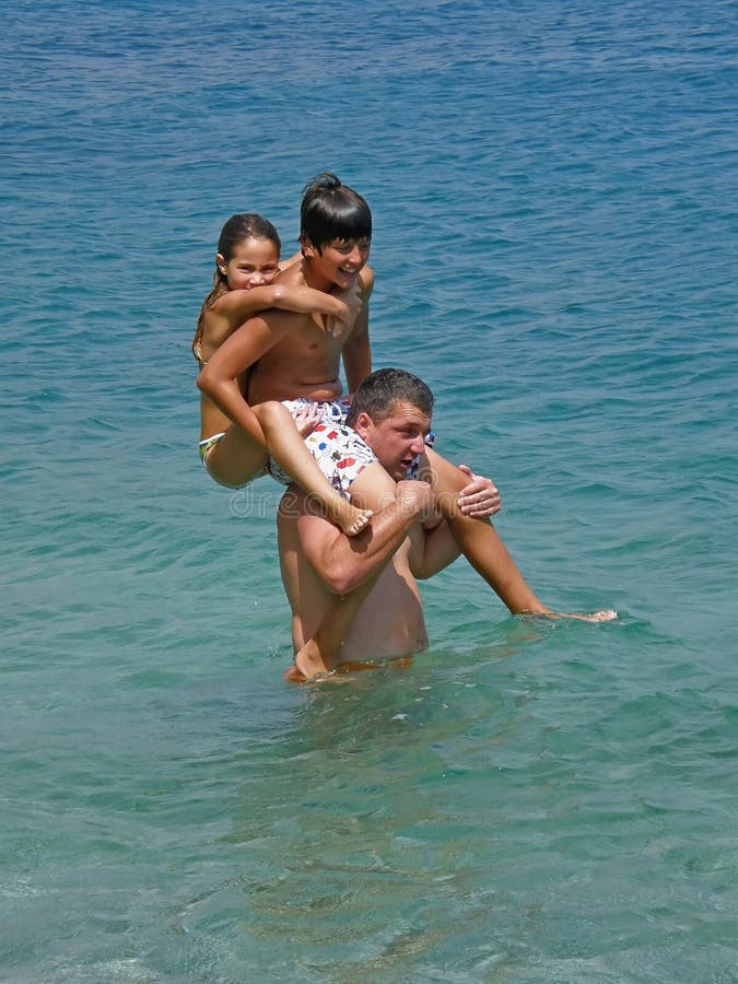 Happy families enjoy in spending summer holidays in Dalmatia (Croatia). The father carried on your shoulders is a son s daughter, (piggy back) and he walks in the blue Adriatic sea. Vertical color photo. Happy families enjoy in spending summer holidays in Dalmatia (Croatia). The father carried on your shoulders is a son s daughter, (piggy back) and he walks in the blue Adriatic sea. Vertical color photo.