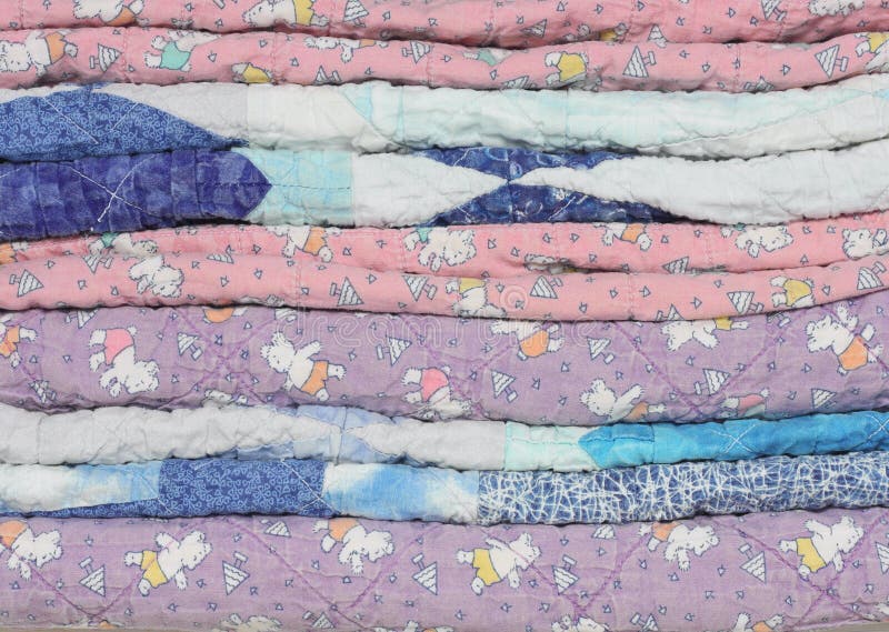 A stack of faded, fraying and worn handmade purple, blue and pink children's quilts. A stack of faded, fraying and worn handmade purple, blue and pink children's quilts.