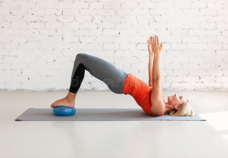 Pilates with small fit ball. Caucasian woman practice gluteal bridge balance on a mat, in loft white studio indoor, selective focus. Workout, fitness, coach, healthy lifestyle concept. Pilates with small fit ball. Caucasian woman practice gluteal bridge balance on a mat, in loft white studio indoor, selective focus. Workout, fitness, coach, healthy lifestyle concept.