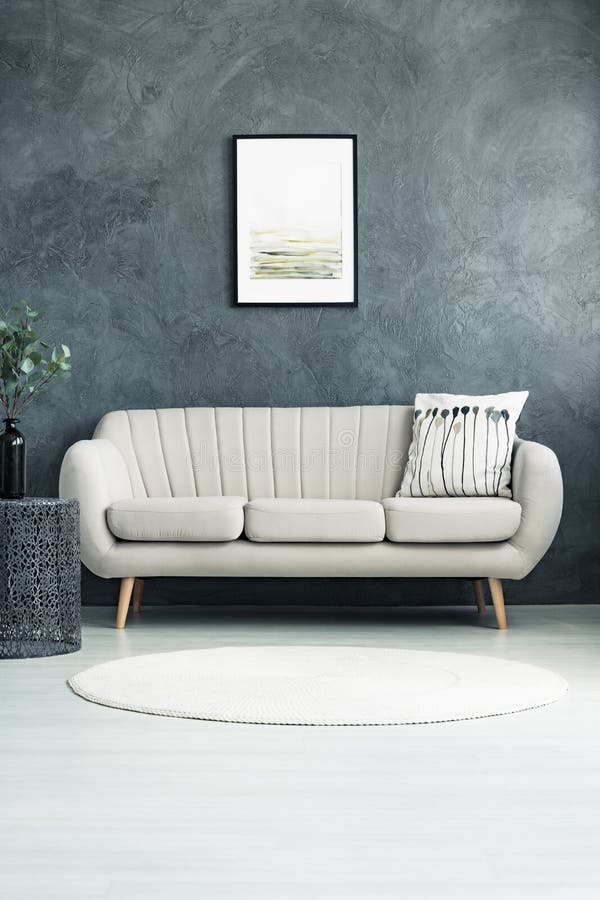 Pillow on beige sofa against concrete wall with poster in living room with white carpet and vase on table