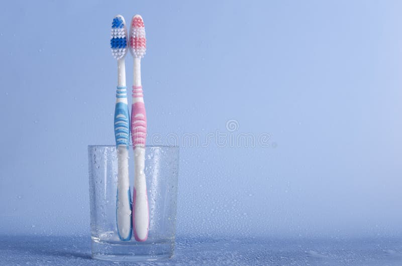 Pink and blue toothbrush in the glass cup on the blue background with water drops